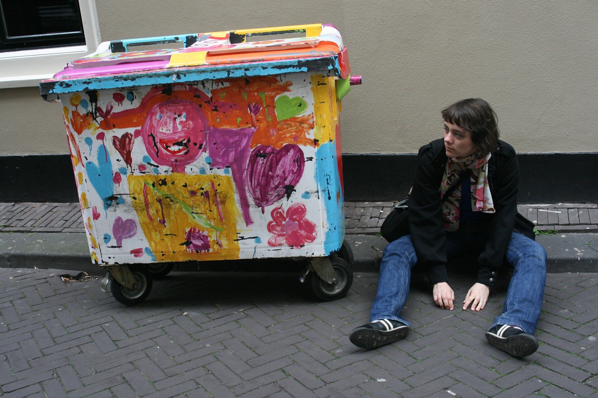 Na ilustracji znajduje się siedząca na krawężniku kobieta. Obok niej stoi kontener na śmieci.