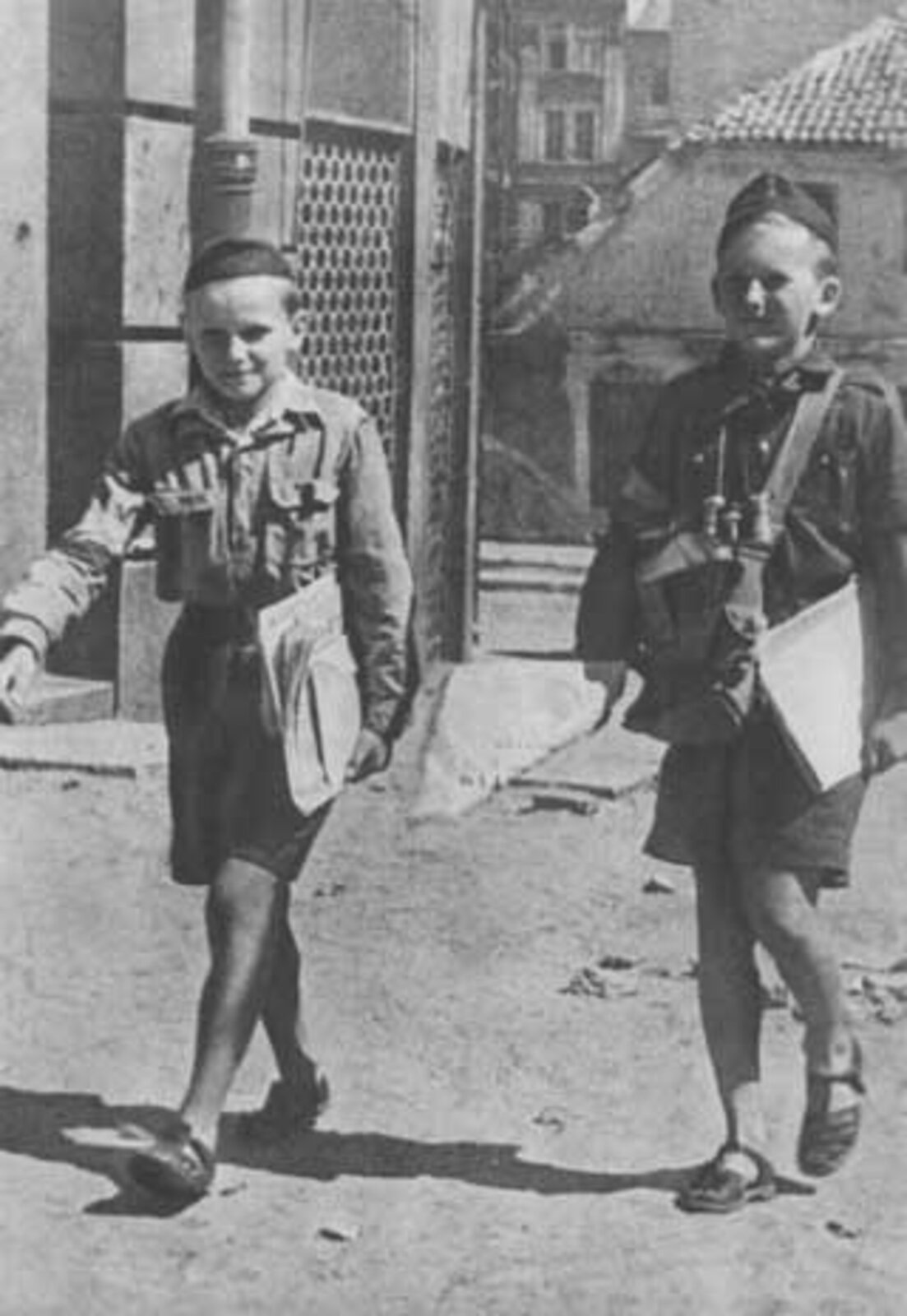 Fotografia czarno-biała przedstawiająca najmłodszych powstańców służących na poczcie. Są to dwaj chłopcy ubrani w mundury. Każdy z nich niesie plik dokumentów i listów do dostarczenia. W tle widoczne są miejskie budynki.