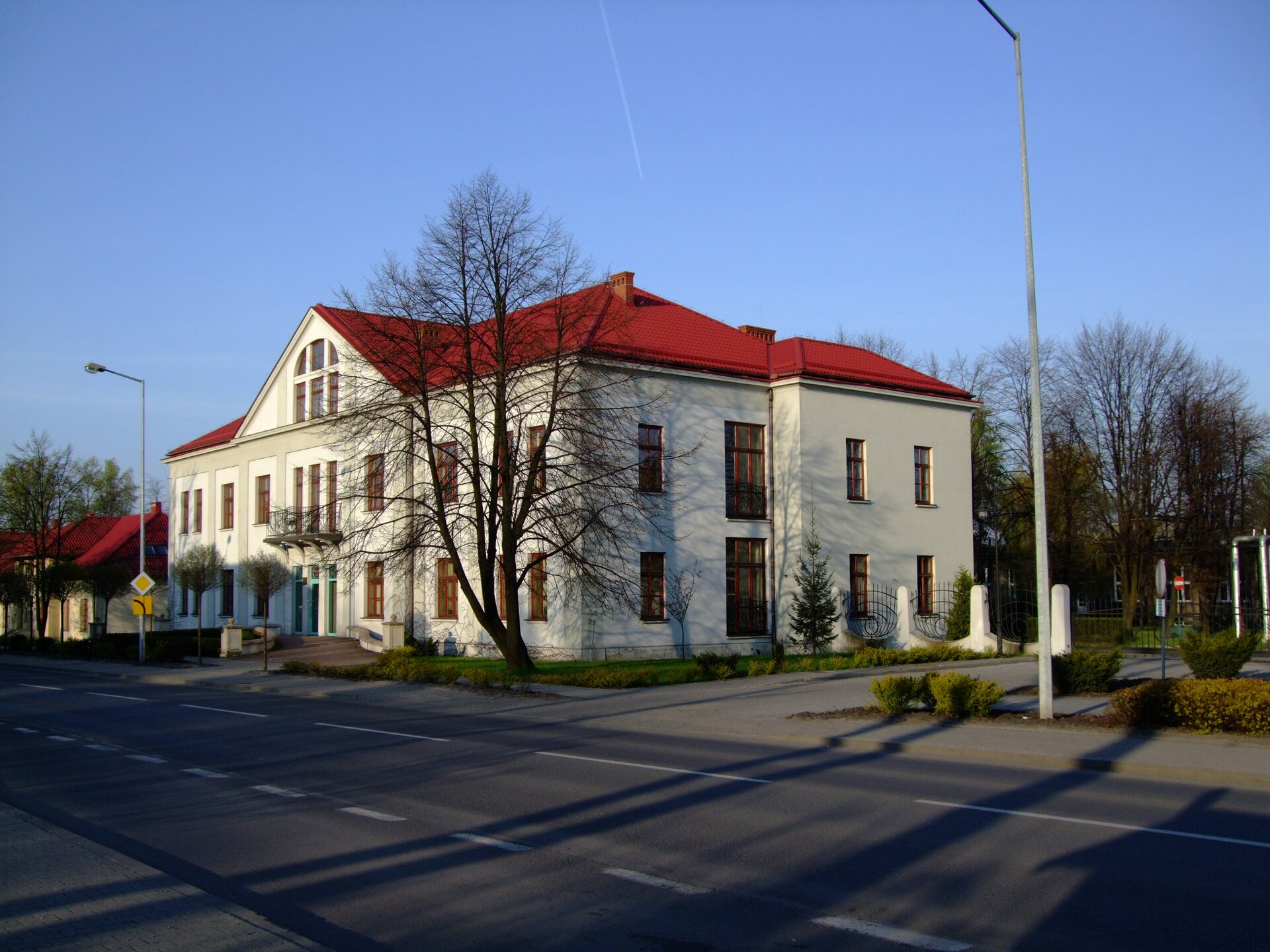 Zdjęcie przedstawia biały budynek z czerwonym dachem znajdujący się przy asfaltowej drodze. Przed nim rośnie drzewo.