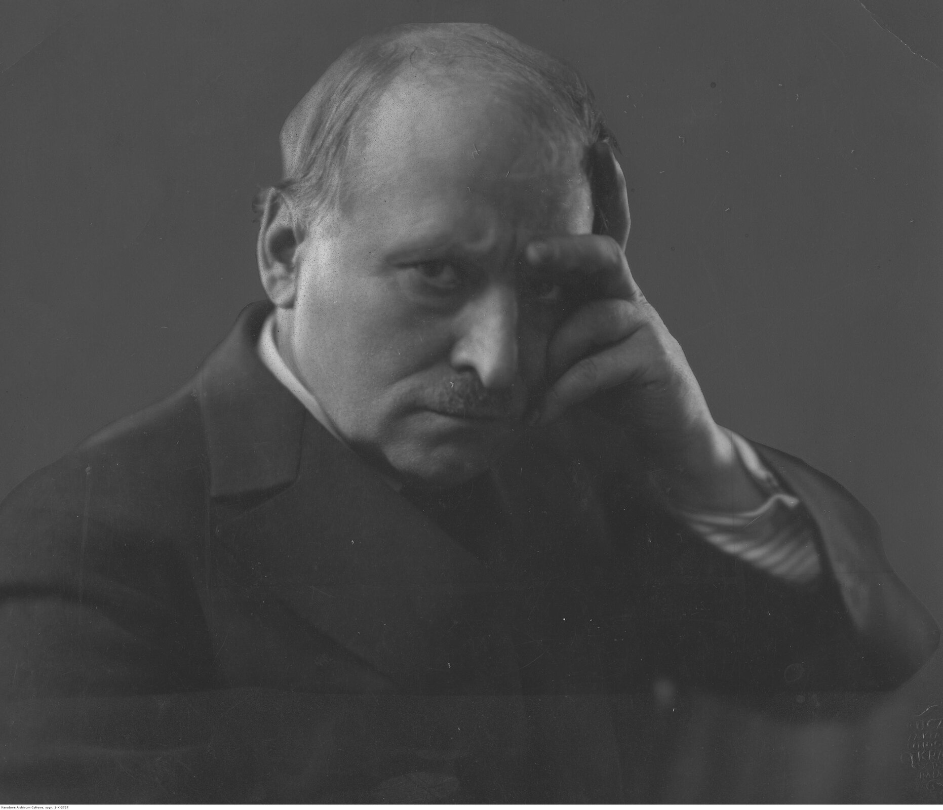 Czarno‑biała fotografia mężczyzny w średnim wieku. Ma on krótkie siwe włosy oraz krótkie wąsy. Ma na sobie czarny płaszcz, spod którego wystaje biała koszula. Ma zaciśnięte usta i zmarszczone brwi. Mężczyzna został uchwycony w dramatycznej pozie z półprofilu – jedną ręką podpiera głowę i patrzy wyrazistym spojrzeniem wprost na widza.