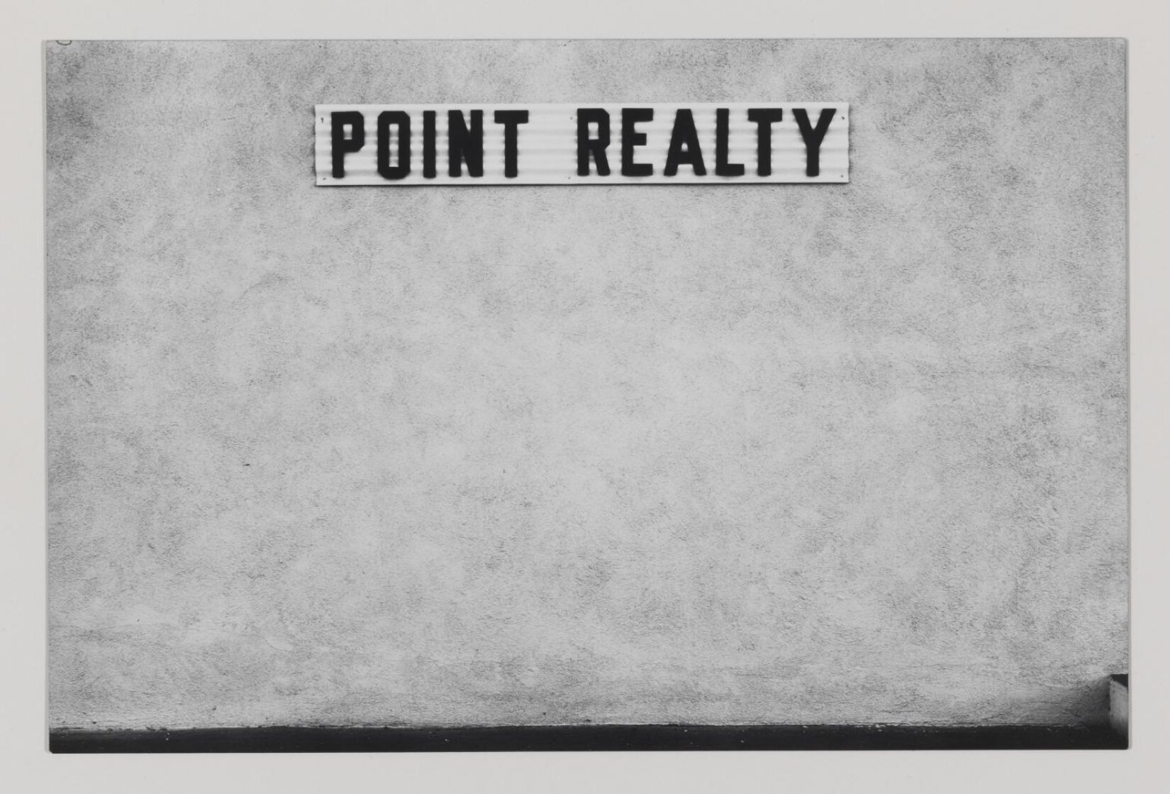 Praca Lewisa Baltza pod tytułem „Dana Point#2” przedstawia fotografię wykonaną w stylu minimalistycznym. Autor zrobił zdjęcie białej ściany, na której znajduje się napis POINT REALTY. Na ścianie widnieją ciemniejsze plamy, co sprawia, że jej struktura nie jest gładka. Wzdłuż dolnej krawędzi zdjęcia przebiega czarny pasek. Praca jest czarnobiała.