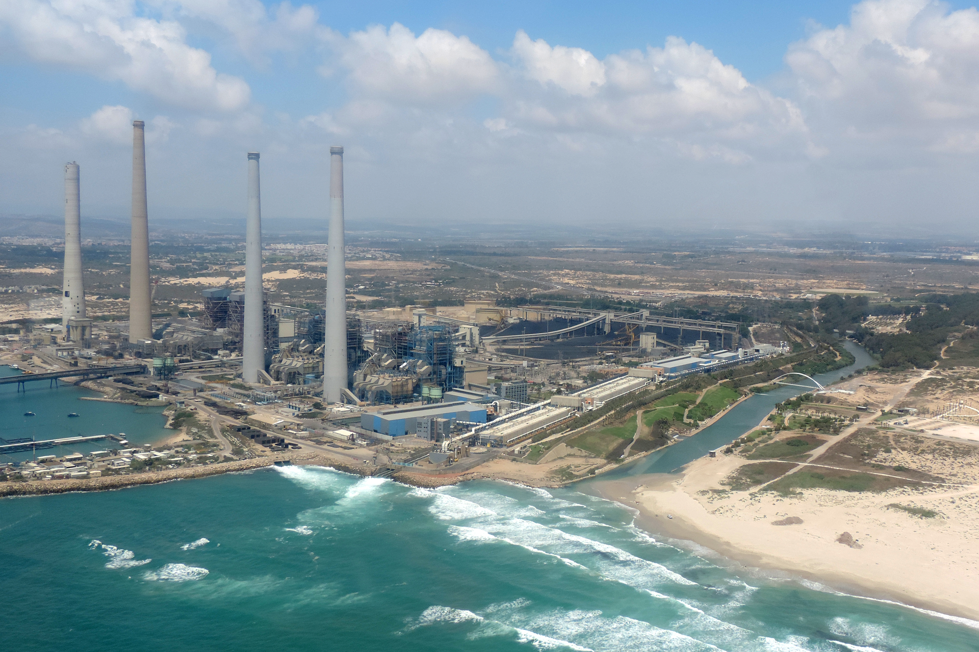 Grafika przedstawia zdjęcie największej na świecie stacji odsalania wody w Izraelu, która znajduje się przy brzegu morza. Na zdjęciu widoczna jest piaszczysta plaża, niebieskie morze z białymi niskimi falami. Przy brzegu są 4 wysokie szare okrągłe kominy. Za kominami są niskie szare budynki stacji.