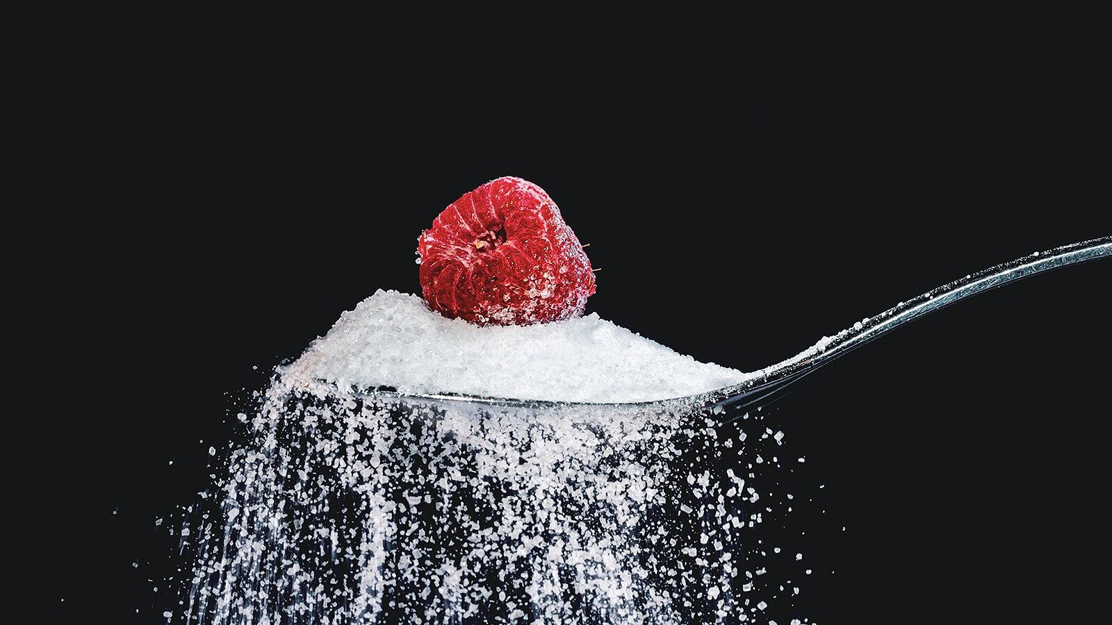 Zdjęcie przedstawia srebrną łyżeczkę z trzonkiem po prawej stronie, na której widoczne są zsypujące się kryształy białego cukru. Na górce cukru na łyżeczce widoczna jest czerwona malina. Całość przedstawiona jest na czarnym tle.