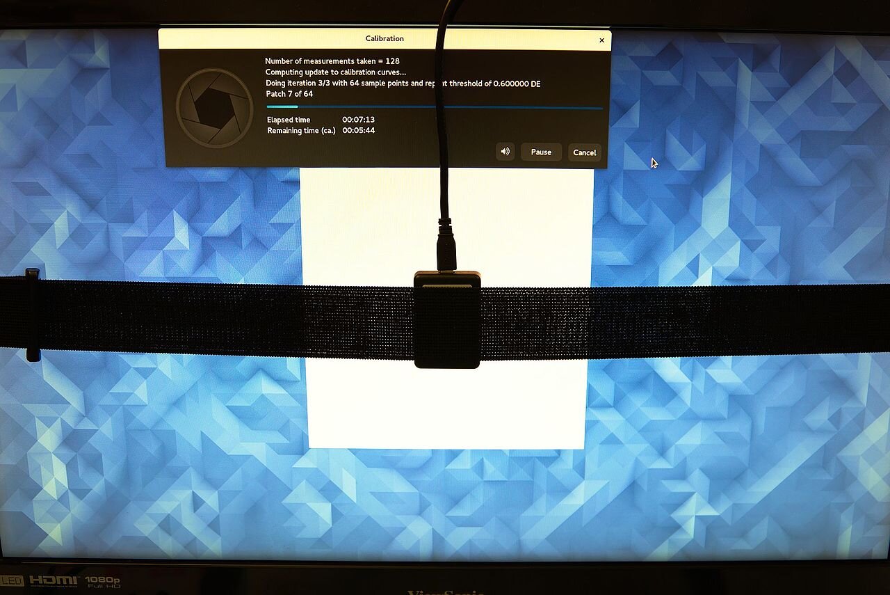 Zdjęcie przedstawia kolorymetr, który jest umieszczony na monitorze, zabezpieczony taśmą mocującą i podłączony kablem do komputera.