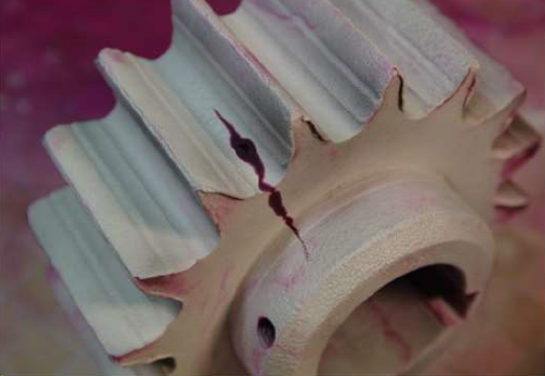 Na zdjęciu znajduje się uszkodzony przedmiot, w którym została wykryta nieciągłość materiału wskutek badania penetracyjnego. Metalowy obiekt posiada widoczne wgłębienie, które jest zabarwione fioletowym płynem.