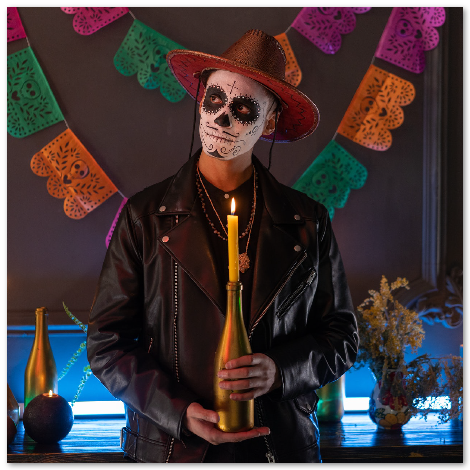 Zdjęcie przedstawia mężczyznę w skórzanej kurtce i kapeluszu kowbojskim. W dłoniach trzyma zapaloną świeczkę. Twarz ma umalowaną w kolorach czarnych i białych, w stylu ozdobnej czaszki.