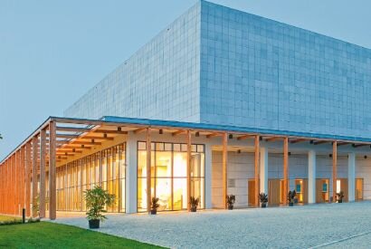 Fotografia przedstawia budynek stojący wśród pól. Jego bryła jest nowoczesna, prosta, centralną jego część stanowi biały, prostokątny budynek bez okien. Otaczają go filary podtrzymujące płaskie zadaszenie. Z tyłu budynku widoczne trzy prostokątne dziedzińce również otoczone płaskim dachem wspartym na filarach. W tle zabudowania Gospodarcze. Fotografia została zrobiona o zachodzie słońca stąd też różowe zabarwienie ciemniejącego nieba.