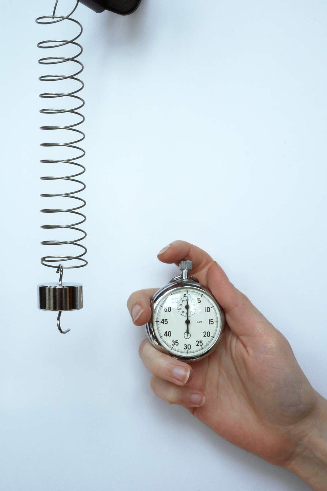Zdjęcie przedstawia ciężarek w postaci metalowego płaskiego walca z haczykami w podstawach. Ciężarek jest zawieszony na długiej sprężynie. Obok widać stoper trzymany przez dłoń. Na stoperze wskazówka wskazuje 0. Tło jasnoniebieskie.