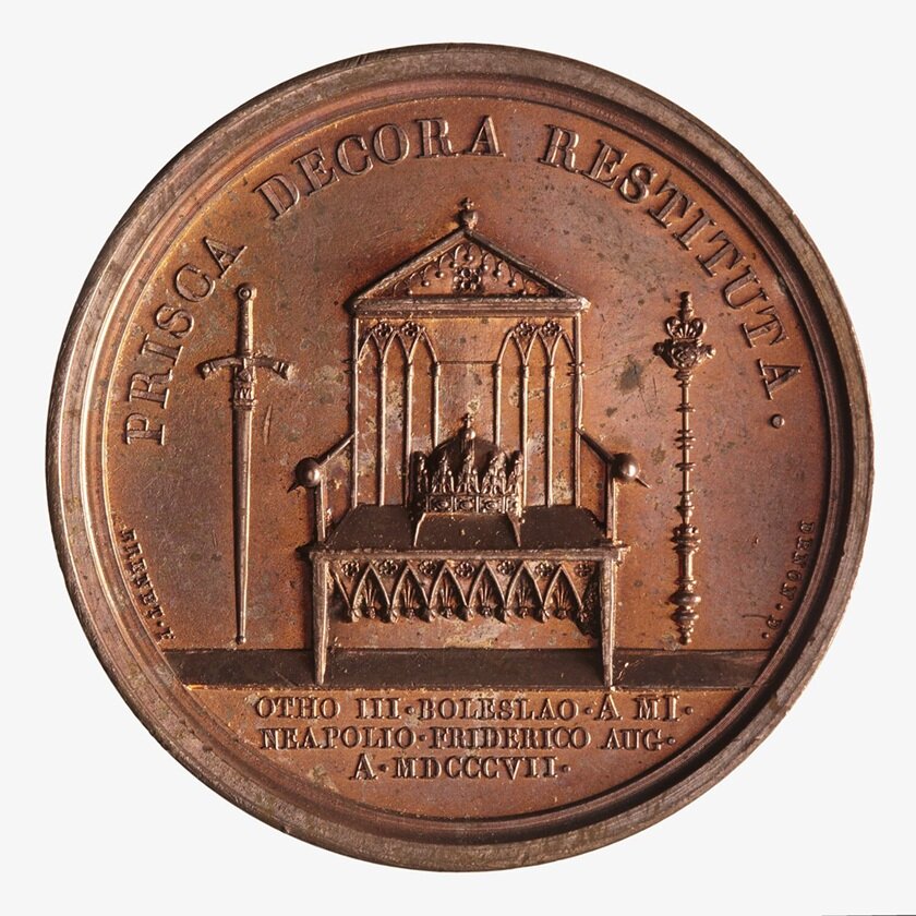 Zdjęcie przedstawia rewers medalu. Pośrodku znajduje się tron, na którym leży królewska korona. Po prawej stronie tronu stoi pionowo miecz, po lewej berło królewskie.