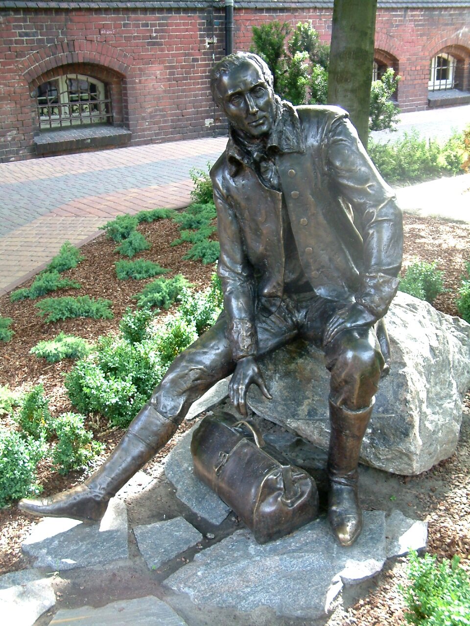 Zdjęcie przedstawia pomnik dojrzałego mężczyzny, który siedzi na kamieniu. Sięga prawą ręką do torby, która leży pod nim. Mężczyzna ma pociągłą twarz, ma krótkie włosy i ubrany jest w surdut. Wokół pomnika są niskie nasadzenia. W tle widoczny jest chodnik oraz ceglany budynek.
