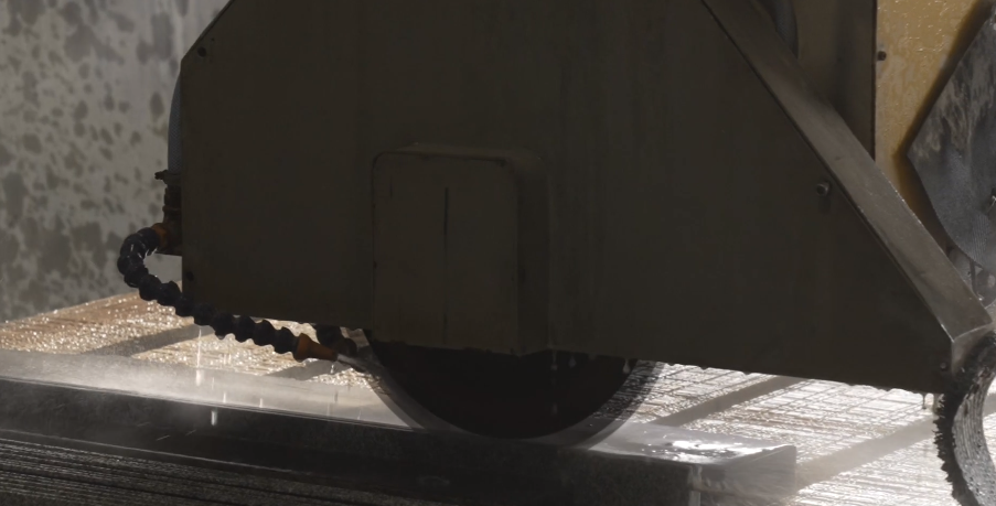 Zdjęcie piły mostowej. Tarcza tnąca zawieszona jest nad stołem roboczym z materiałem kamiennym. Widoczny jest wężyk doprowadzający wodę w miejsce cięcia.