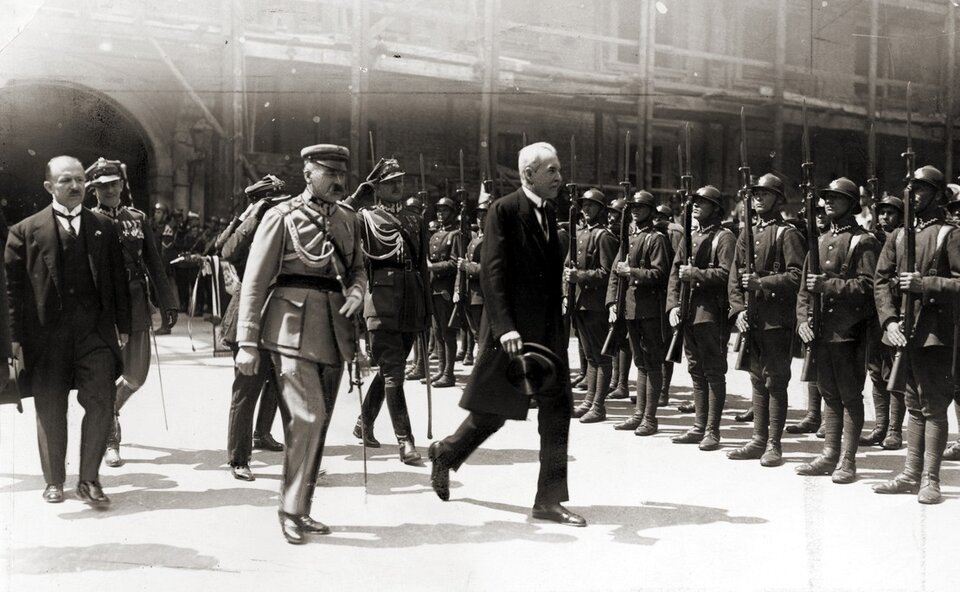 Zdjęcie przedstawia idących mężczyzn naprzeciwko stojących na baczność żołnierzy z bronią. Na czele idą dwaj mężczyźni: mężczyzna w mundurze wojskowym oraz mężczyzna w garniturze. 