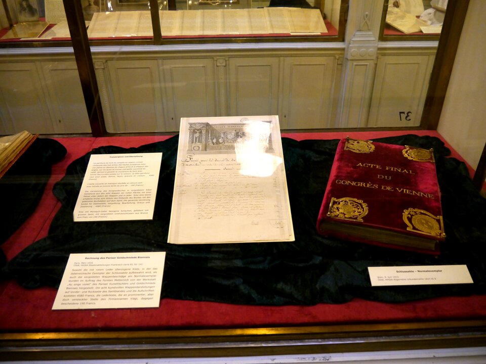 Fotografia przedstawia szklaną gablotę, w której na aksamitnym kawałku materiału leżą dwie zapisane kartki i obok książka w eleganckiej oprawie ze złotymi zdobieniami.