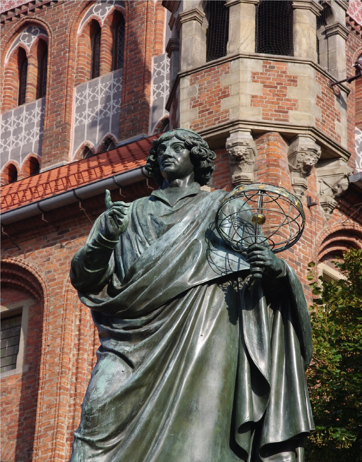 Ilustracja przedstawia posąg Mikołaja Kopernika w Toruniu. Rzeźba ukazuje stojącego mężczyznę w średnim wieku z półdługimi, falowanymi włosami. Postać okryta jest pofałdowaną peleryną. W lewej dłoni trzyma ażurowy model układu słonecznego. Prawą rękę z wysuniętym do góry, wskazującym palcem ma zgiętą w łokciu i lekko uniesioną. Odlana z brązu, ciemno‑zielona rzeźba ustawiona jest na tle wykonanej z czerwonej cegły, barokowej fasady.