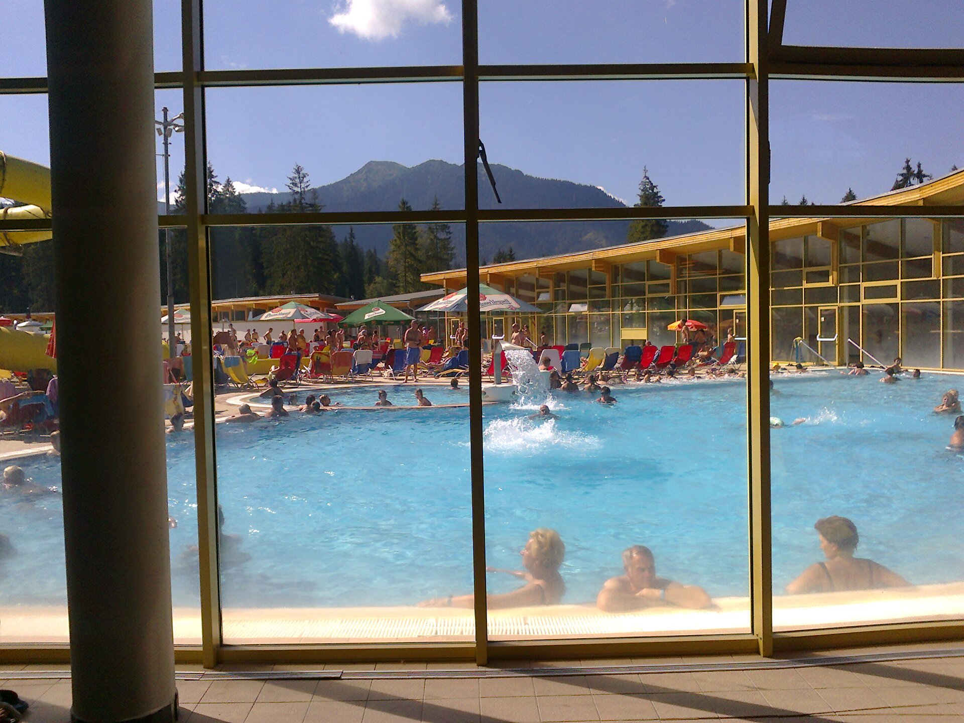 Na zdjęciu basen zewnętrzny, w którym kąpią się dorośli i dzieci. Basen znajduje się za szklaną ścianą. Na brzegu basenu widoczna jest infrastruktura wykorzystywana przez turystów. Są to parasole, oraz zadaszona strefa wypoczynkowa. Nad basenem widoczny jest tłum turystów. W tle basenu góry. 