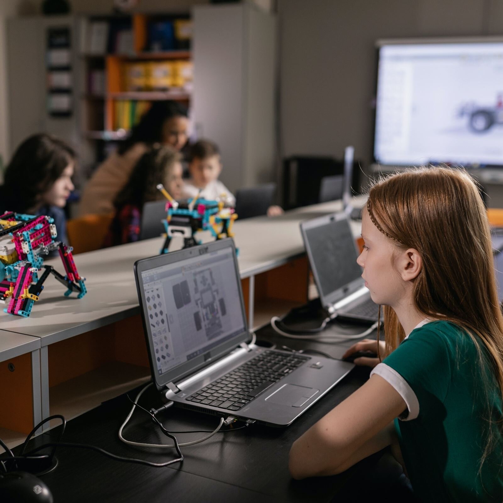 Zdjęcie przedstawia dziewczynkę w wieku około 11 lat, siedzącą przy biurku na którym stoi laptop. W tle siedzą inne dzieci, również przy komputerach.