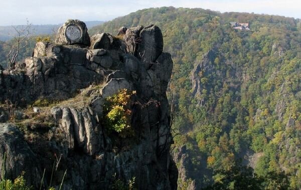 Zdjęcie przedstawia góry Harz. Na pierwszym planie, po lewej stronie widoczna jest skała z nieregularnymi głazami i prawie pionowym zboczem. W tle góry porośnięte drzewami.