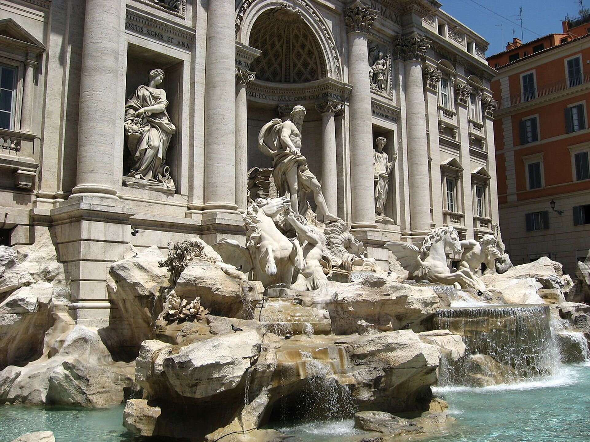 Kolorowa ilustracja przedstawia słynną rzymską fontannę Di Trevi. Na fotografii ukazane jest ujęcie z prawej strony fontanny. Jest to monumentalna budowla składająca się z bogato dekorowanej ściany oraz kamiennych elementów, na których umieszczono rzeźby. W centrum fontanny stoi Neptun w powozie zaprzężonym w hipokampy. Bóg przedstawiony jest w pozycji stojącej. Ma umięśnione ciało i rozwianą przez wiatr szatę. Spogląda w dół powozu. Za boskim rydwanem, w niszach ściennych, znajdują się kobiece postaci będące alegoriami Zdrowia i Obfitości. Ze skalnych elementów spływa do basenu woda.