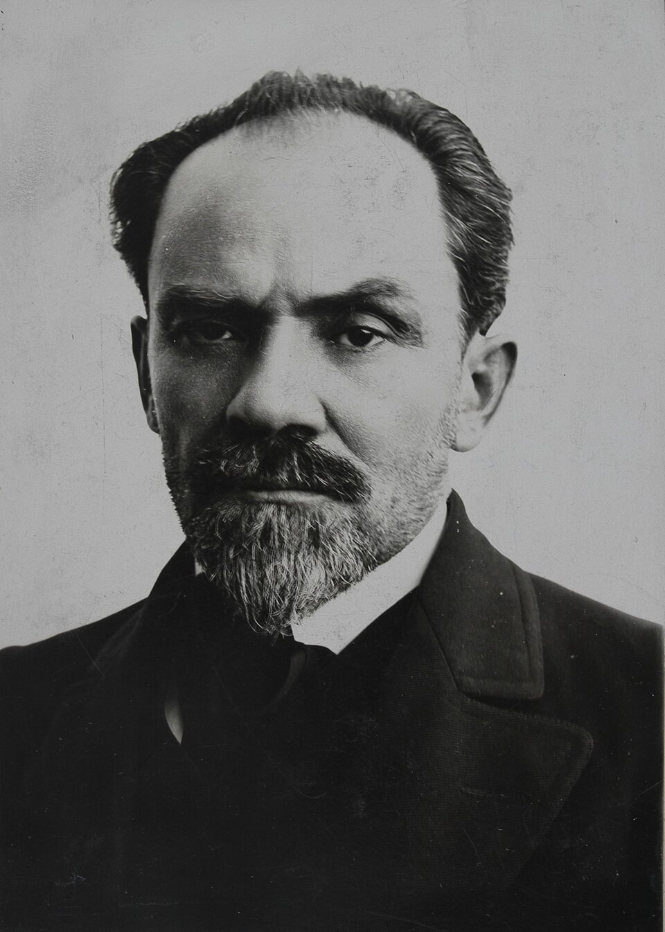 Zdjęcie przedstawia mężczyznę w średnim wieku o poważnym wyrazie twarzy. Ma krótkie, lekko cofnięte włosy i gęstą brodę z wąsami. Jego brwi są gęste i lekko zmarszczone. Ubrany jest w elegancki, ciemny garnitur z kołnierzykiem, koszulę. 