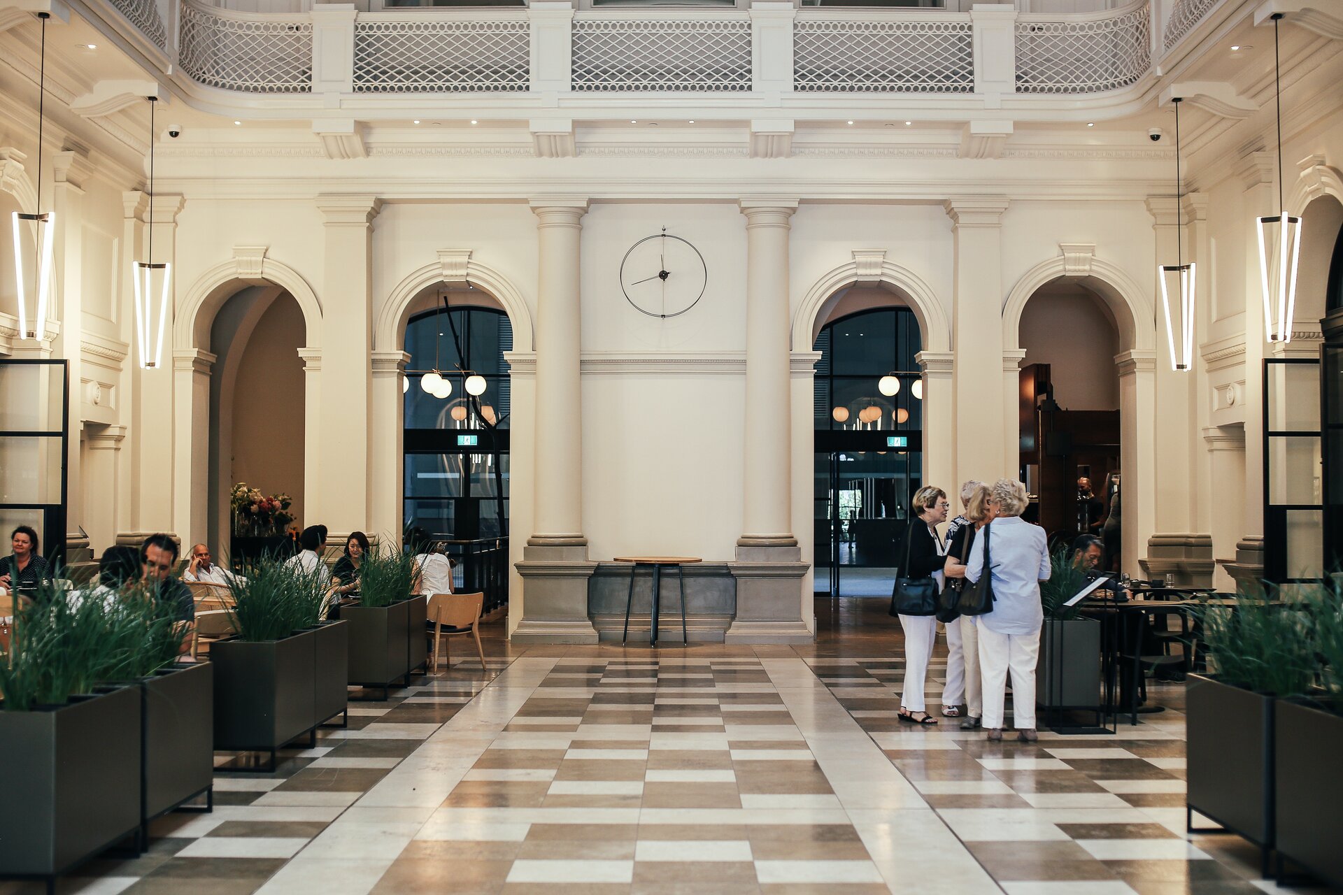 Zdjęcie przedstawia hall przykładowego hotelu. Widoczne jest monumentalne wnętrze z białymi ścianami i półkolumnami w tle oraz marmurową podłogą z ciemnych i jasnych płyt. Po lewej stronie widoczne są wydzielone strefy do siedzenia z elementami roślinnymi. Po prawej trzy kobiety stojące przy stendzie.