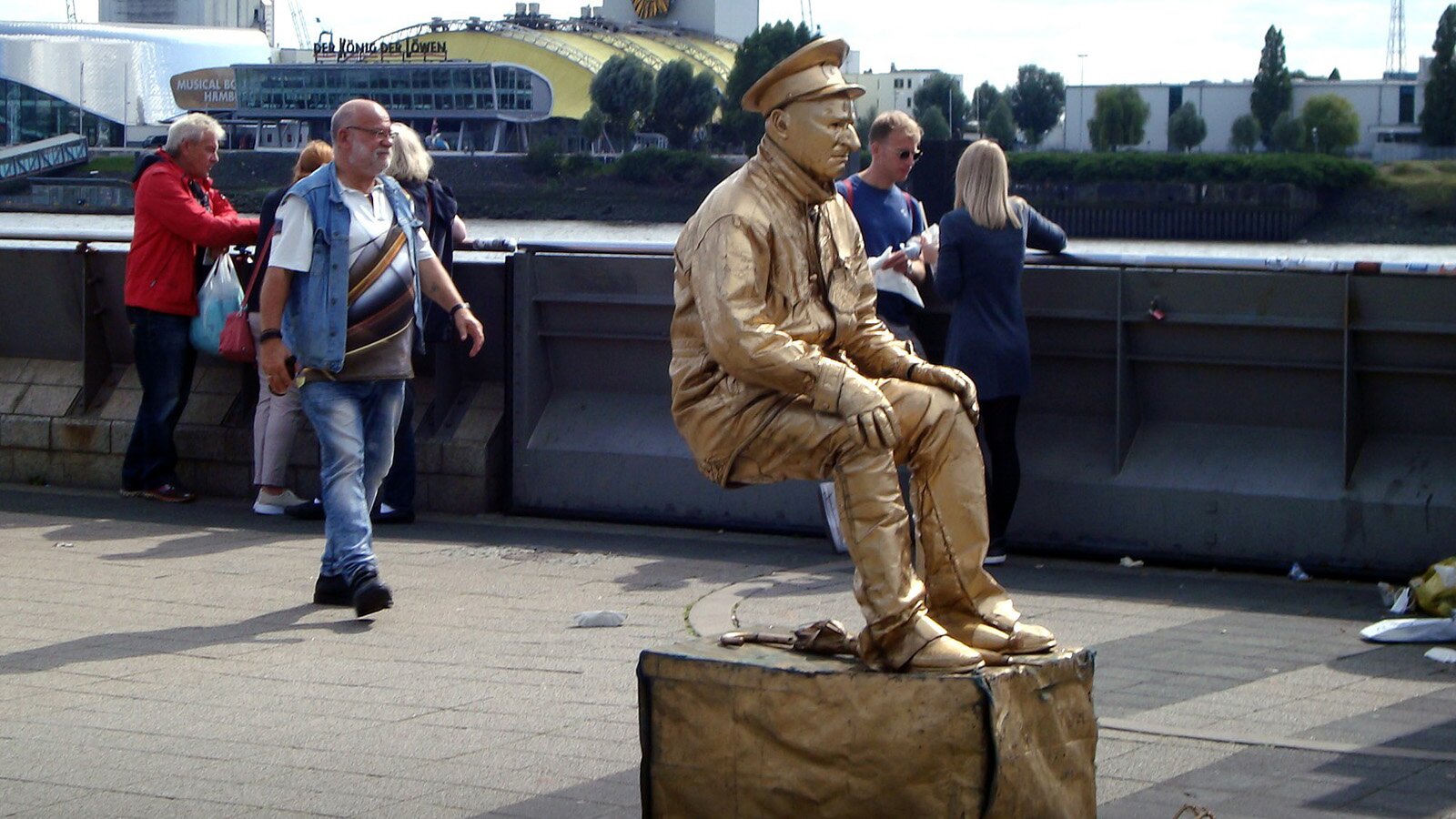 Zdjęcie przedstawia mężczyznę ubranego oraz pomalowanego całego na złoto. Mężczyzna jest ukazany w pozycji siedzącej, lecz bez żadnego podparcia. Postać siedzi w powietrzu.