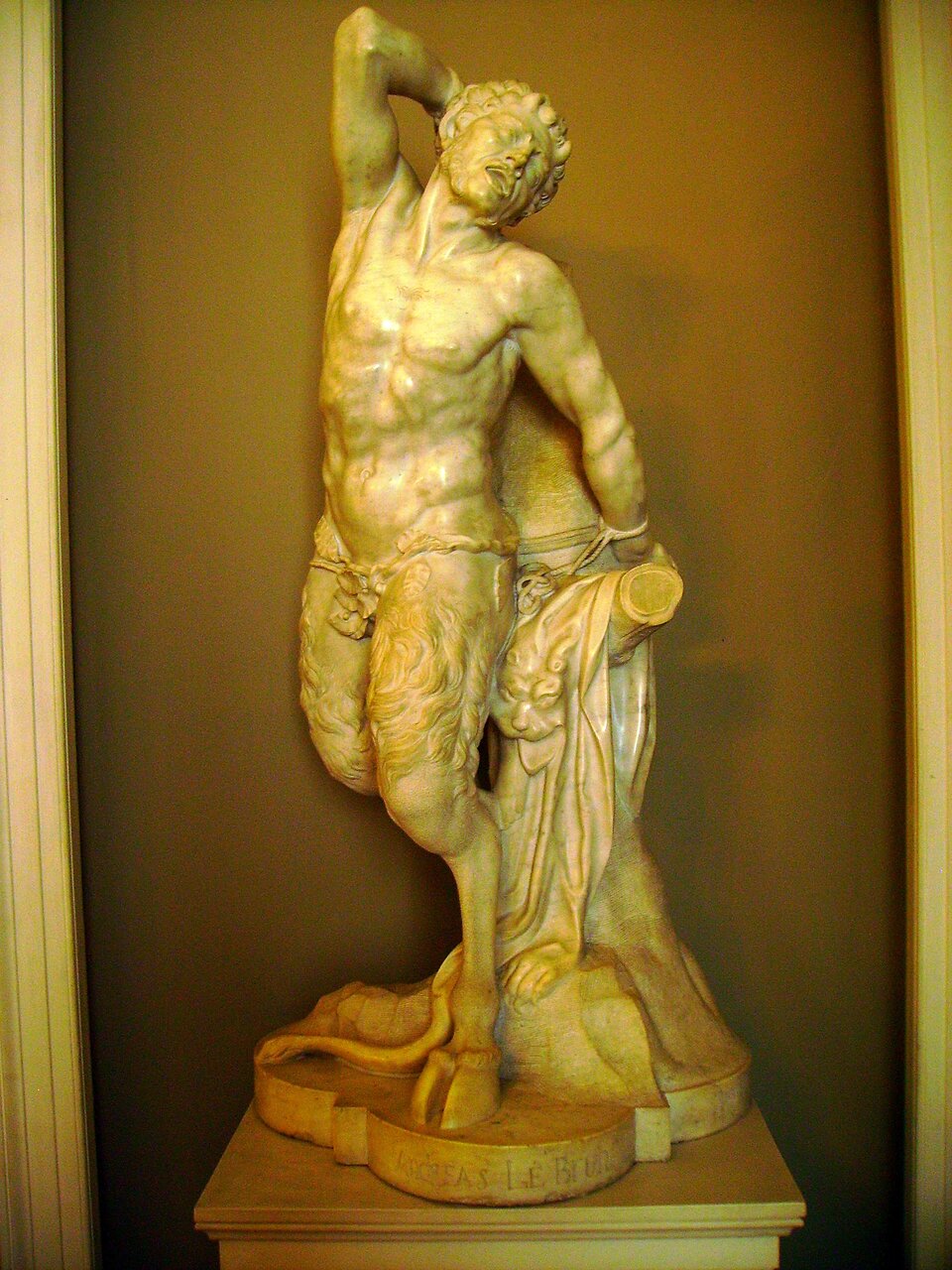 Fotografia przedstawia rzeźbę André Le Bruna pod tytułem „Marsjasz". Satyr Marsjasz, to nagi, szczupły mężczyzna z nogami kozła i małymi różkami na głowie. Na biodrach ma przepaskę z liści osłaniającą genitalia. Stoi przy konarze drzewa, do którego jest za lewą rękę przywiązany. Głowę ma odgiętą w tył, usta wykrzywia grymas. Prawą rękę, zgiętą w łokciu unosi do góry. Satyr opiera się o konar ściętego drzewa, który jest przykryty skórą lwa. Cała rzeźba jest ustawiona na podstawie z wyrytym nazwiskiem autora.