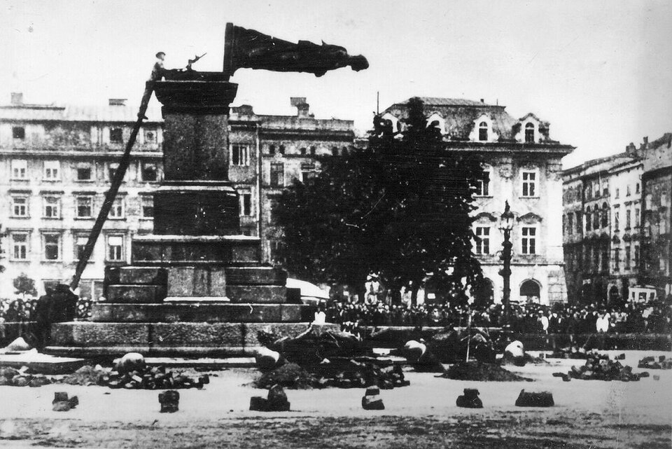 Zdjęcie przedstawia plac w środku miasta, dookoła którego zebrali się ludzie. Na środku stoi postument, do którego przystawiono drabinę. Na drabinie stoi człowiek. Zdjęcie uchwyciło moment, w którym pomnik Adama Mickiewicza znajduje się w w chwili upadku na bruk. Postać zwisa poziomo do ulicy, za chwilę upadnie głową w dół. Dookoła leżą rozrzucone szczątki otoczenia pomnika.