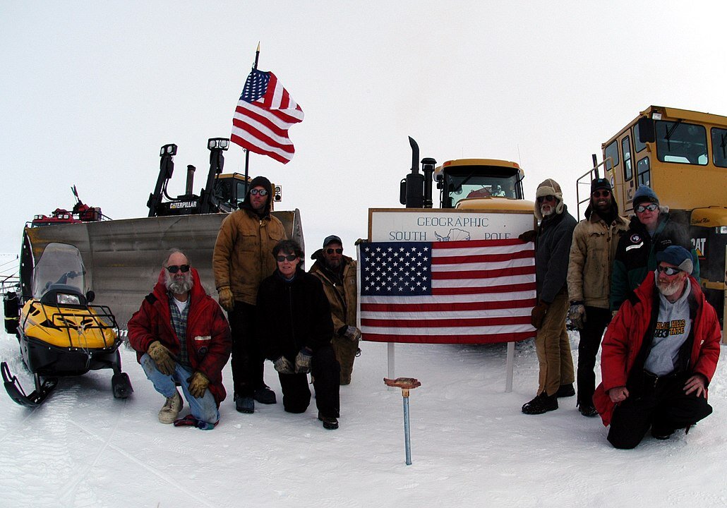 Zdjęcie przedstawia grupę osób zebranych wokół tablicy informacyjnej umieszczonej na biegunie południowym. Ubrani w ciepłe ubrania, dwójka z nich trzyma flagę Stanów Zjednoczonych. Za nimi skuter śnieżny i ciężki sprzęt.