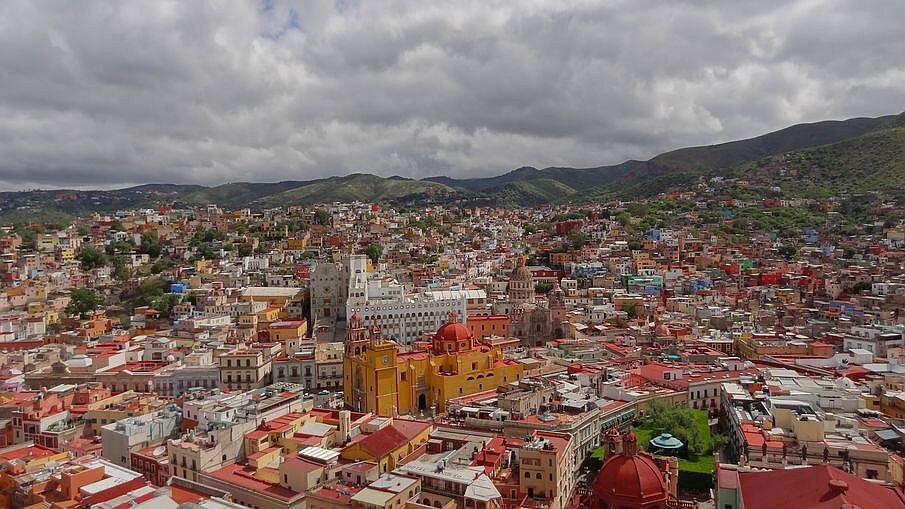 Zdjęcie przedstawia duże miasto. Zabudowa jest gęsta. Dominują niskie budynki. W centralnej części fotografii jest masywny obiekt zwieńczony kopułą, a za nim wyższy biały budynek i kolejny zakończony kopułą. Większość budynków ma płaskie, pomarańczowe dachy. W tle jest pasmo górskie.
