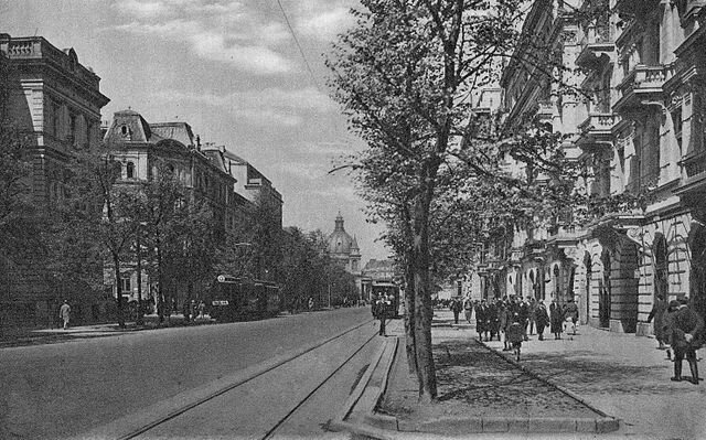 Czarno‑biała fotografia przedstawia Aleje Ujazdowskie przed 1939 rokiem. Widok przedstawia ulicę, po obu jej stronach znajdują się kamienice dwupiętrowe, chodniki, po których idą ludzie oraz tory tramwajowe, z jadącymi tramwajami. W tle widać kopułę kościoła placu Trzech Krzyży.