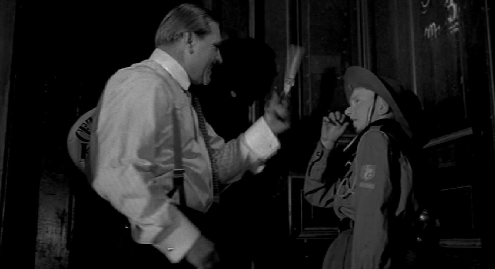 Zdjęcie przedstawia nastoletniego chłopca w stroju harcerskim i stojącego naprzeciw niego mężczyznę w koszuli i spodniach. Mężczyzna trzyma przed twarzą chłopca nożyczki. 