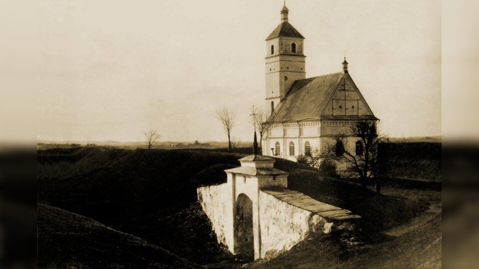 Zdjęcie przedstawia wysoki wał z ziemi z niewielką murowaną bramą. Na drugim planie widoczna jest niewielka cerkiew z wysoką wieżą.