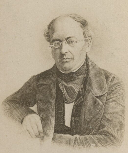 Ilustracja przedstawia portret mężczyzny. Jest łysiejący, ma krótkie włosy po bokach. Nosi okulary w małych, okrągłych oprawkach. Ma lekko zarysowane bokobrody. Na jego twarzy widoczny jest drugi podbródek. Mężczyzna ubrany jest w surdut.