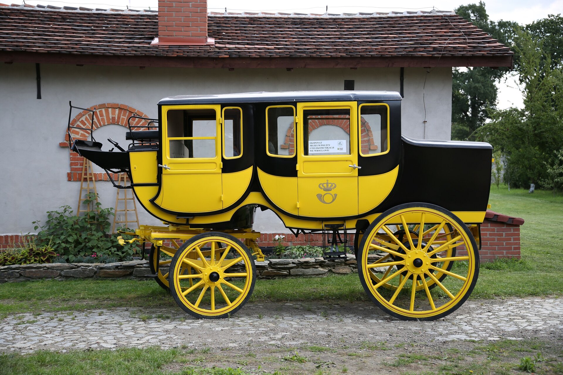 Ilustracja przedstawiająca dawny powóz pocztowy, z dwoma większymi kołami z przodu i dwoma mniejszymi z tyłu. Na podwoziu osadzona jest kabina i dwie pary drzwi do niej, w środku są miejsca siedzące. Powóz stoi na wybrukowanej drodze. W tle za powozem znajduje się budynek urzędu pocztowego. 