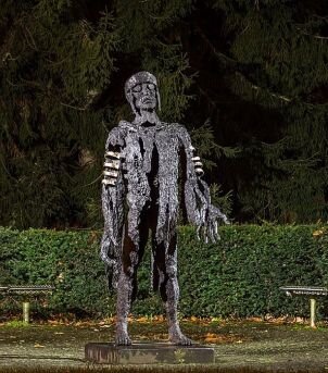 Zdjęcie przedstawia metalową rzeźbę mężczyzny. Ma on przecięty od klatki piersiowej w dół, wyciągnięte do przodu ręce z dużymi dłońmi, głowę z założonym hełmem odchyla lekko w tył. Rzeźbę otacza ciemnozielony żywopłot. 