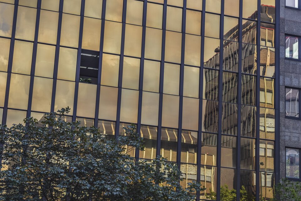 Fotografia przedstawia fragment nowoczesnego, przeszklonego budynku. Ukazane są duże okna. Jedno z nich jest otwarte. W szybach odbija się zachmurzone niebo oraz dachy innych budynków.