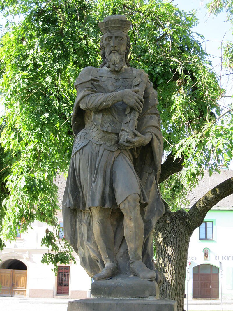 Na zdjęciu znajduje się kamienny pomnik Świętopełka. Jest on ubrany w szatę i lekką zbroję. Ma długie włosy i brodę. W rękach trzyma zwój dokumentów. Za pomnikiem rośnie duże drzewo. 