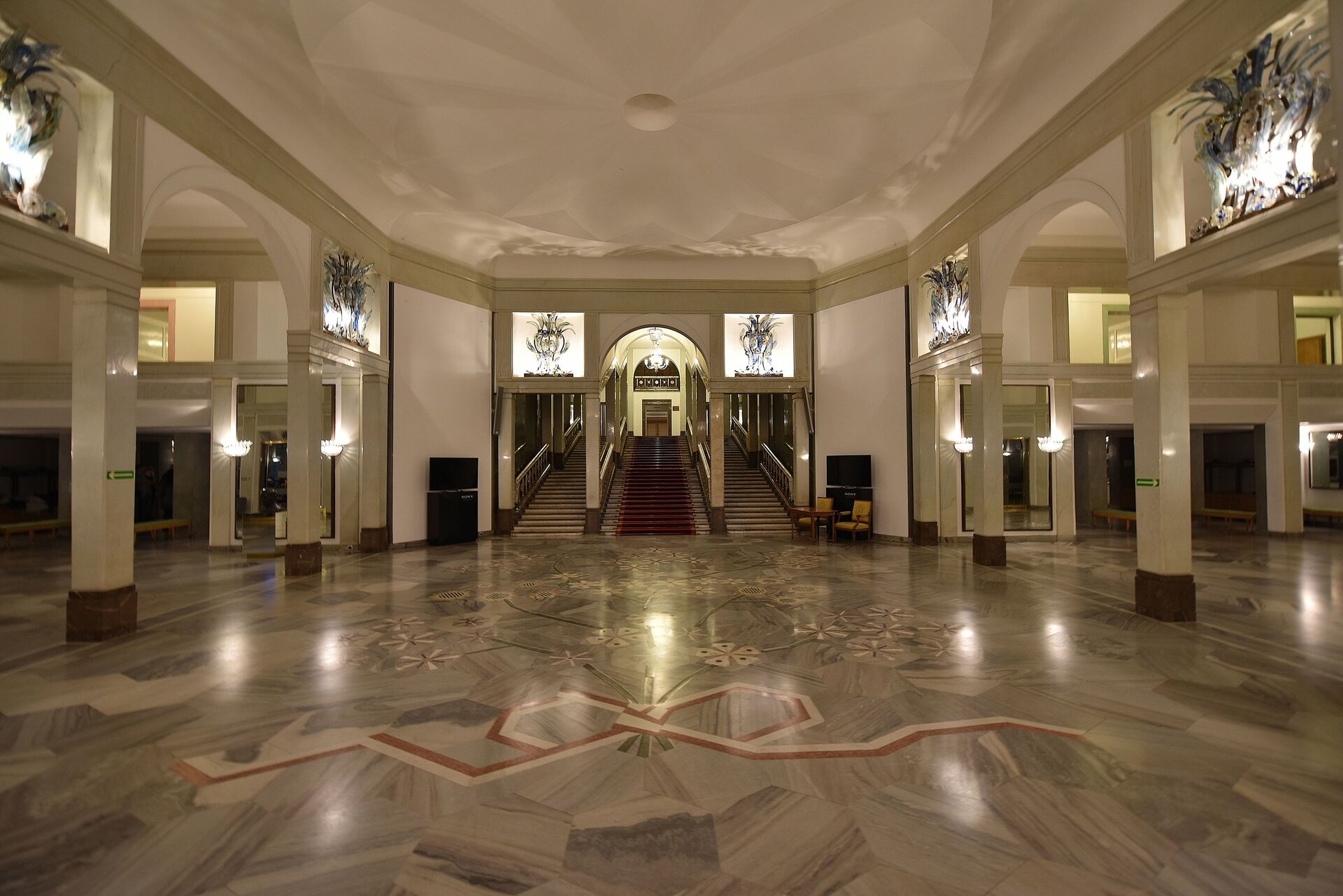 Fotografia przedstawia bogato zdobione wnętrze budynku. Na pierwszym planie na podłodze znajduje się bukiet kwiatów w beżowo‑szarej tonacji, gałązki bukietu przewiązane są biało‑czerwoną wstęgą zawiązaną na kokardę. Cała podłoga wykonana z kamiennych płyt w kształcie plastra miodu. Po prawej i lewej stronie holu beżowe kolumny zwieńczone łukami. Pomiędzy łukami, nad kolumnami znajdują się dekoracyjne szklane żyrandole z ciemnego szkła. Na wprost trzy wejścia na schody, środkowe wejście zwieńczone łukiem. Przed schodami cztery kolumny. Środkowe schody wyłożone dywanem w kolorze bordowym. Na szycie schodów widoczne są drzwi. Sufit holu zdobi prosta rozeta w kolorze kremowym.