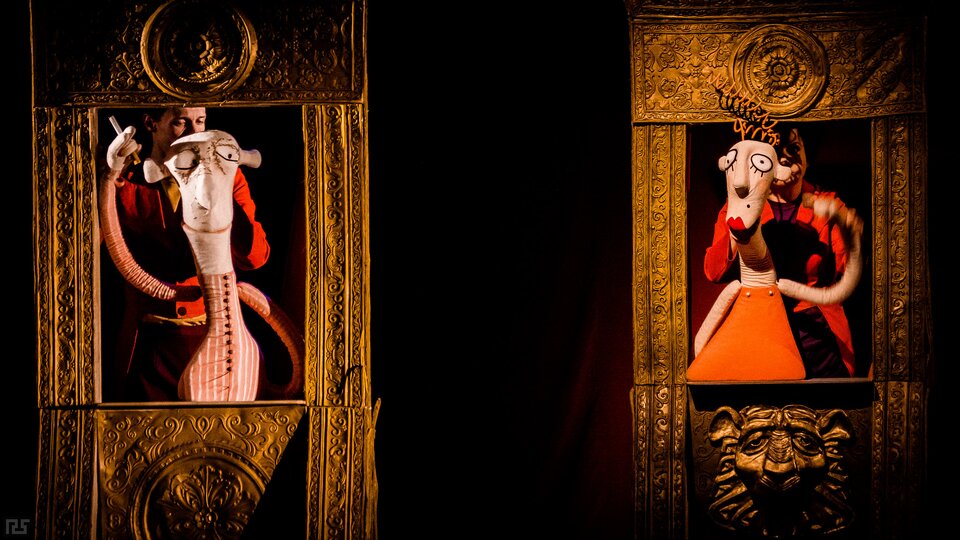 Fotografia przedstawia scenę teatralną. Stoją tu dwie wąskie konstrukcje przypominające ramy luster lub obrazów. Stoją za nimi lalki wielkości dorosłych ludzi. Po lewej stronie ukazana jest postać mężczyzny. Ma dużą głowę o trójkątnej twarzy. Jest łysy. Ma duże, okrągłe oczy, długi nos, niewielkie uszy i szerokie usta. Unosi prawą rękę, trzymając papieros. Jest ubrany w marynarkę. Po prawej stronie ukazana jest postać kobiety. Jej głowa jest duża, a twarz trójkątna. Ma na głowie kilka sprężynek wyobrażających włosy. Jej oczy są  duże, okrągłe, nos długi, uszy niewielkie, a usta uszminkowane. Ma niewielki pieprzyk nad wargą, po lewej stronie twarzy. Unosi lewą rękę. Jest ubrana w prostą sukienkę.