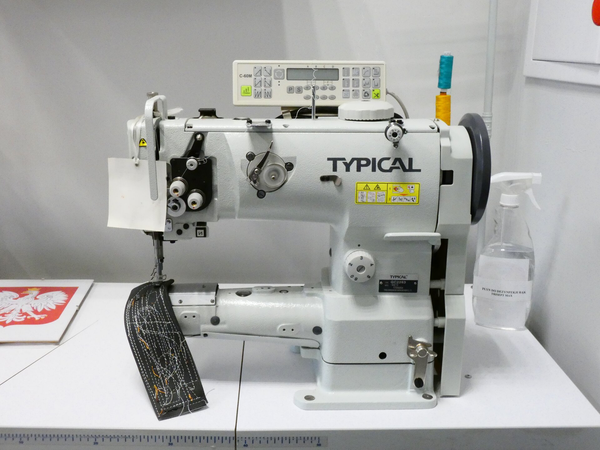 Zdjęcie przedstawia białą, dwupoziomową maszynę. Na dolnym panelu leży kawałek materiału, górny panel jej szerszy, na górze znajdują się przyciski sterowania. 