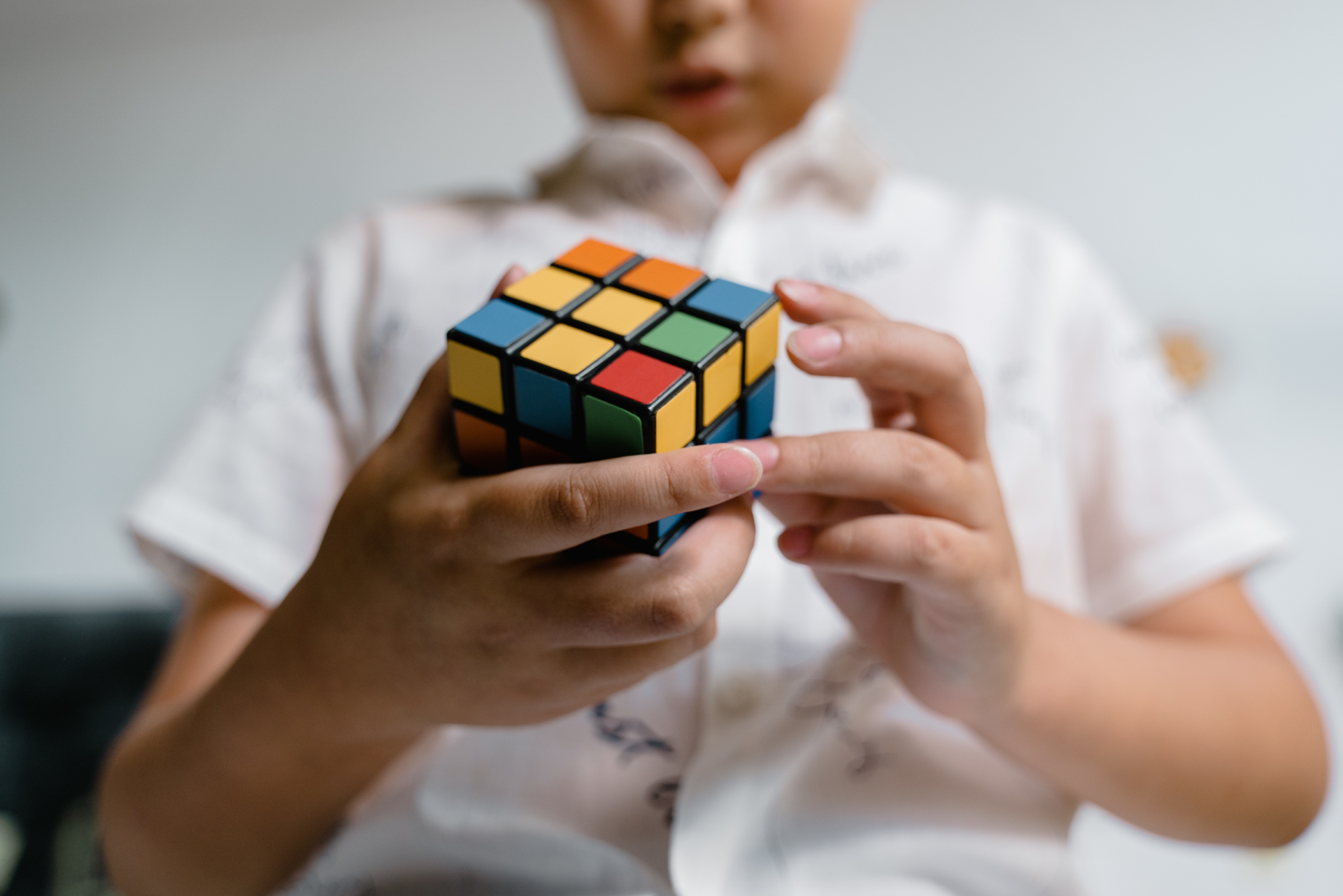 Zdjęcie przedstawia chłopca, w ujęciu od pasa do wysokości nosa, który w dłoniach trzyma kostkę Rubika. 
