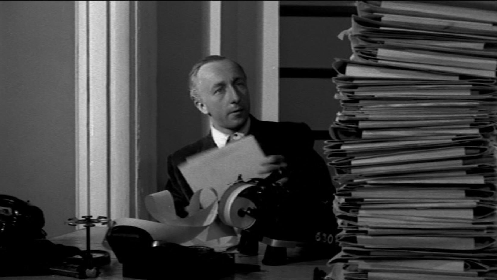 Zdjęcie przedstawia głównego bohatera siedzącego przy biurku. Na biurku piętrzy się ogromny stos teczek z papierami.