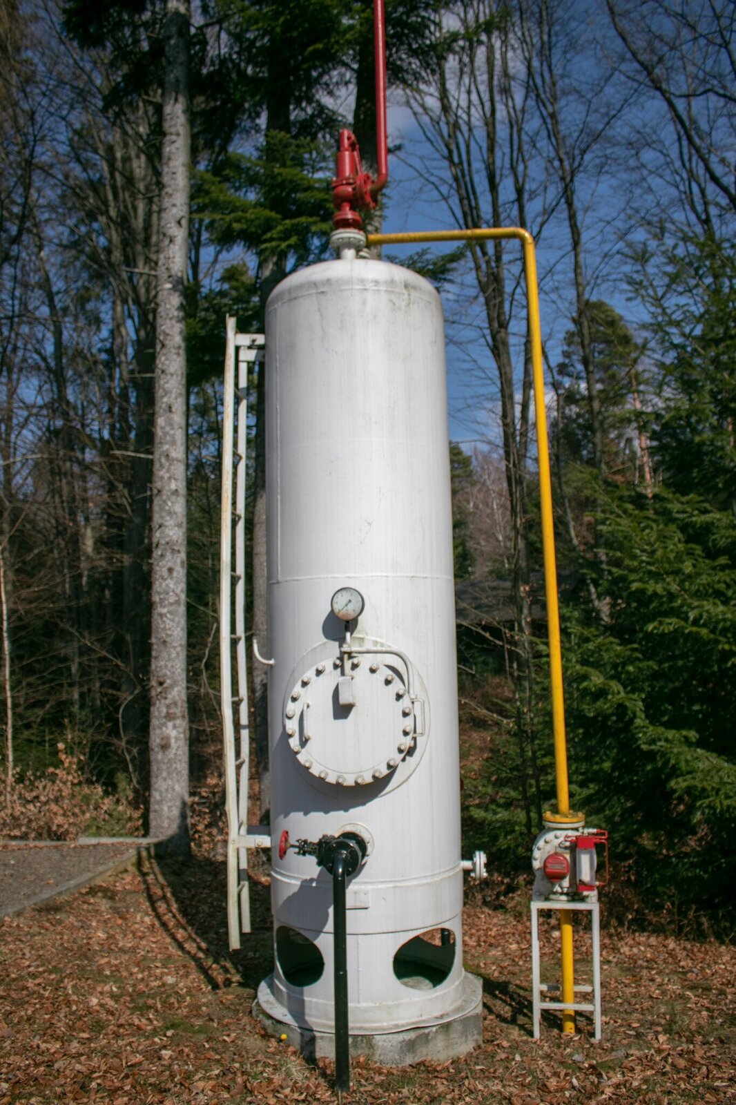 Na zdjęciu widać stalowy separator dwufazowy pionowy typu Rdziesięć łamane na tysiąc, który wykorzystywany jest do rozdziału fazy gazowej od ciekłej. U góry separatora znajduje się czerwony zawór bezpieczeństwa w kształcie odwróconej fajki skierowanej szyjką do góry. Z prawej strony separatora biegnie równolegle do niego cienka stalowa rura. Rura przyłącza się do separatora w miejscu zlokalizowanym pod zaworem bezpieczeństwa. Rura ta wykorzystywana jest do wyprowadzenia gazu. U podstawy rury znajduje się czerwony gazomierz z ekranem ciekłokrystalicznym, który wykorzystywany jest do pomiaru przepływającego gazu. Na dole separatora znajduje się przyłączona do niego czarna cienka rura która zakończona jest od strony separatora czerwonym kurkiem. Jest to zawór spustowy. Powyżej przyłącza rury na separatorze zamontowany jest okrągły manometr, który za pomocą wskazówki pokazuje wartość ciśnienie w zbiorniku. Za separatorem znajdują się drzewa iglaste.