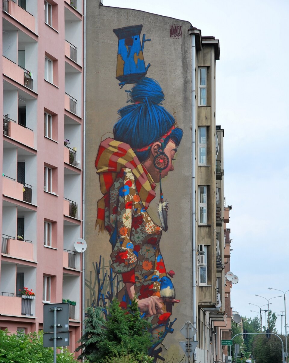 Ilustracja przedstawia mural na szarej ścianie bloku mieszkalnego. Mural przedstawia kobietę w bluzce w kolorowe kwiaty, pod szyją ma szalik w żółto‑czerwoną kratę. Włosy upięte w kok są niebieskiego koloru. W uszach ma kolczyki w kształcie dużego koła z dopiętym na nitce piórkiem ptasim. Kobieta stoi bokiem do widza. Nad jej głową namalowany jest karmnik dla ptaków w kolorze niebieskożółtym.