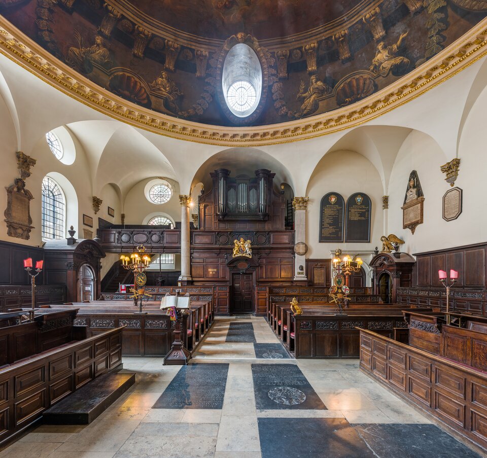 Wnętrze kościoła z drewnianymi ławami i galerią organową na końcu nawy. Nad przestrzenią znajduje się kopuła z dekoracyjnym malowidłem oraz owalnym oknem wpuszczającym światło. Ściany zdobią tablice pamiątkowe i rzeźbione elementy architektoniczne.