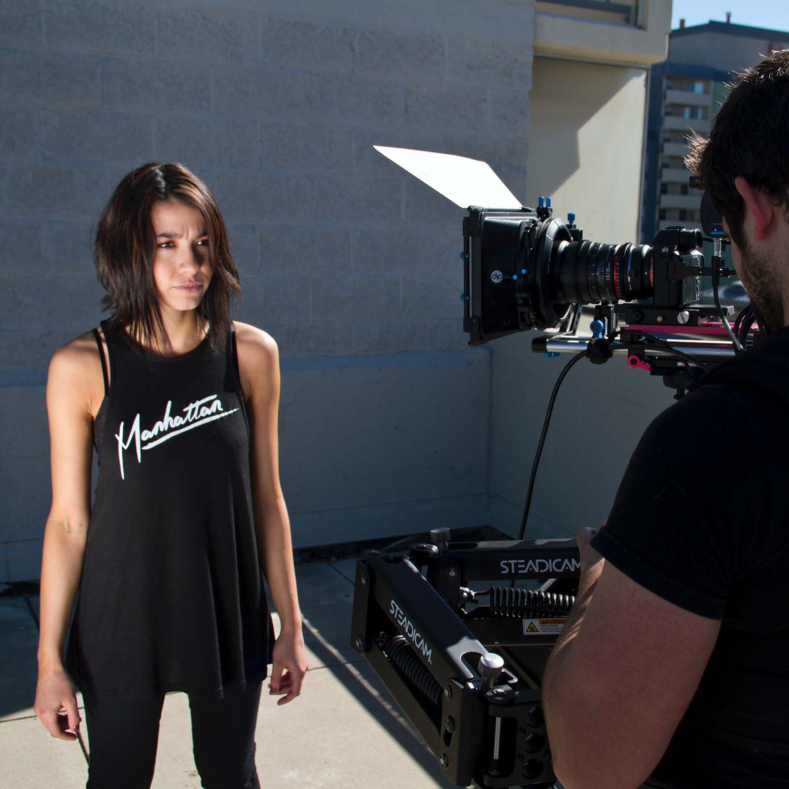 Zdjęcie ukazuje młodą ciemnowłosą kobietę, jej włosy sięgają do ramion. Jest ubrana w czarną koszulkę na ramiączka z białym napisem "Manhattan", oraz czarne długie spodnie. Przed kobietą stoi mężczyzna obsługujący kamerę.