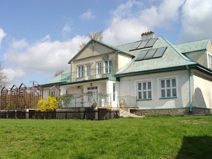 Fotografia przedstawia biały dworek z seledynowym dachem. Wokół trawa. Budynek ma białe ściany, duże okna. Pośrodku, przed wejściem taras, u góry balkon. Na dachu panele fotowoltaiczne. 