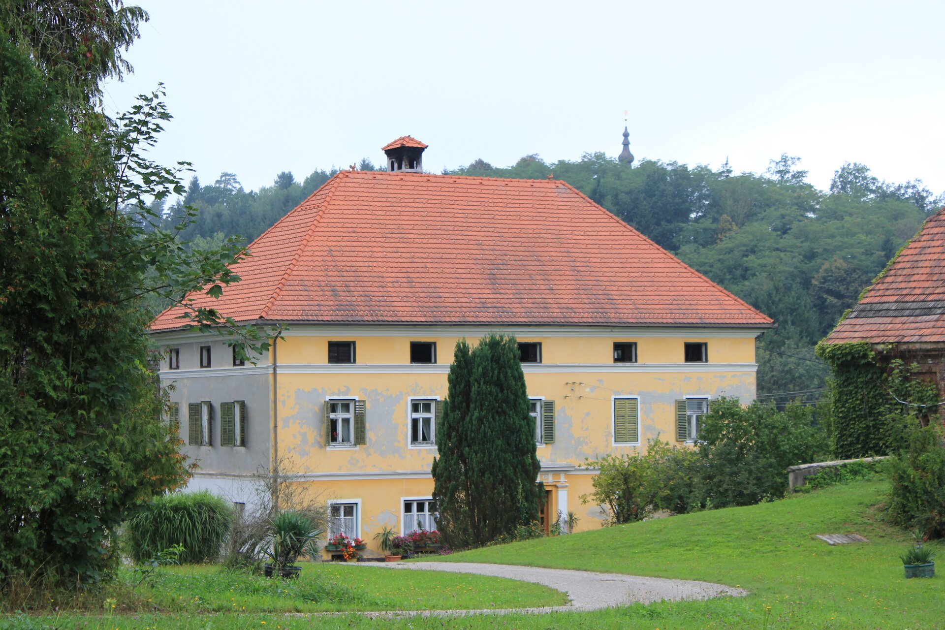 Fotografia przedstawia Preglhof w Obersdorfie, czyli dom rodzinny Weberna. Jest to duży budynek z żółtą elewacją i czerwonym dachem, usytuowany wśród zieleni. Okna maja zielone okiennice. Niebo jest zachmurzone.