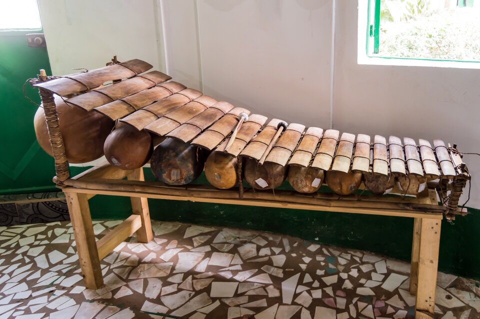 Fotografia przedstawia instrument muzyczny o drewnianej konstrukcji. Podstawa przypomina ławkę o cienkiej desce zamiast siedzenia, następnie z lewej strony obecne są dwa pionowe wystające elementy owinięte materiałem. Pomiędzy nimi umieszczono podłużnie brązowy przedmiot o podłużnym, owalnym kształcie. Następnie, na lewo od niego jest jeszcze osiem podobnych, coraz mniejszych przedmiotów. Ułożono na nich połączone rzemykiem cienkie deseczki, tworzące delikatny łuk. Instrument sfotografowano w pomieszczeniu o brązowo‑kremowej podłodze oraz ze ścianami pomalowanymi na zielono i biało. 