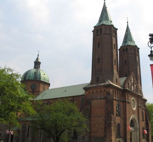 Na zdjęciu znajduje się Bazylika katedralna w Płocku. Jest to ceglany budynek o zielonym dachu. Od frontu posiada ona dwie wieże o spiczastych zakończeniach. Wokół niego rosną drzewa. 